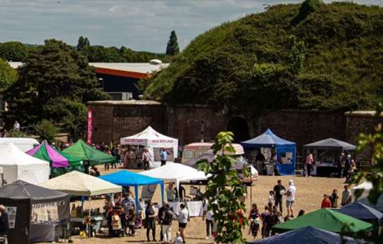 Outdoor craft fair with rows of colourful canopy stalls and visitors in a gravel courtyard beside a red-brick building and trees under a cloudy sky.
