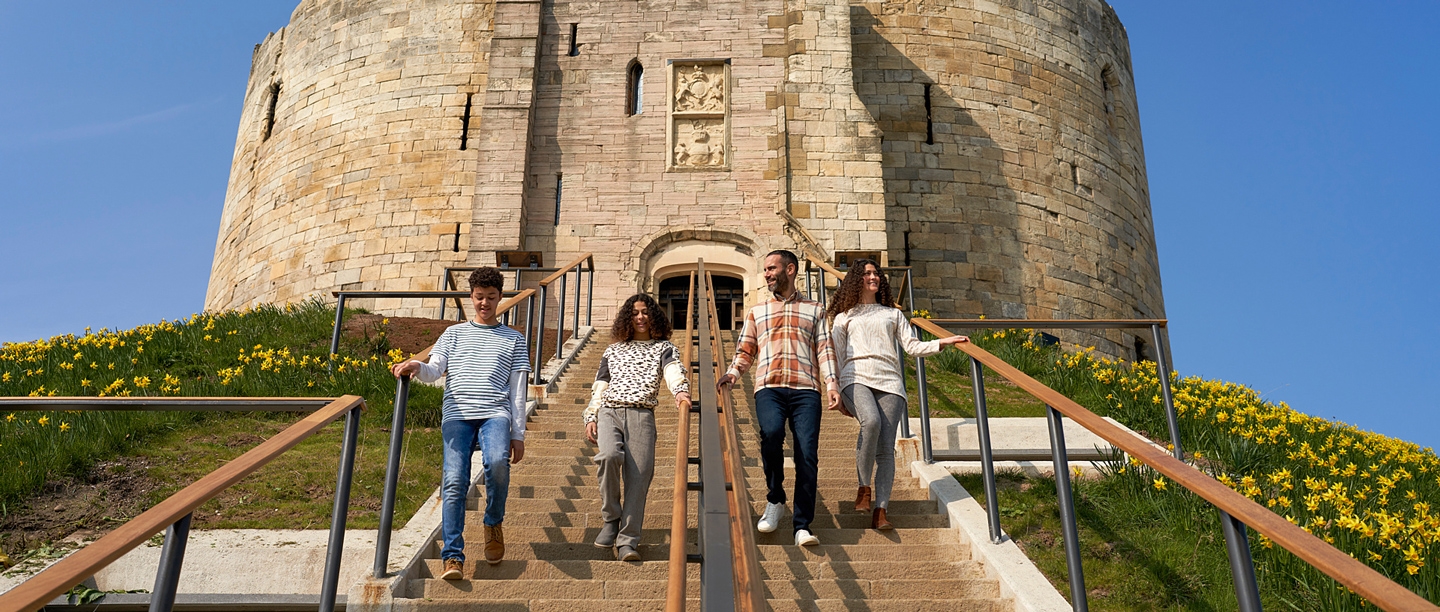 Two adults and two children walk down the steps at Clifford's Tower in York. There are daffodils growing on the mound.