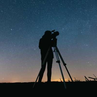 A silhouette of a person using a telescope on a tripod, standing under a clear night sky filled with stars.