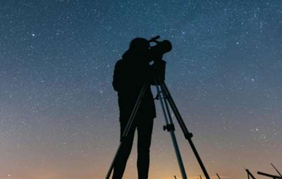 A silhouette of a person using a telescope on a tripod, standing under a clear night sky filled with stars.